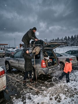 A family packs bags and gear onto their car roof rack in a snowy parking lot, getting ready for a winter road trip.