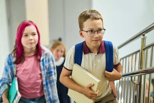 Caucasian schoolboy with glasses holding book and walking up stairs with classmates. Image represents primary education and is suitable for school enrollment campaigns
