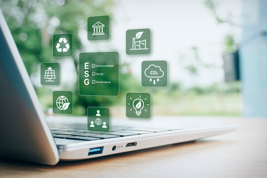Laptop computer on wooden desk with holographic ESG environmental, social, and governance icons, green energy, sustainability, and corporate social responsibility business concept.