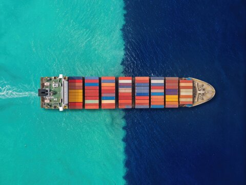 Top-down aerial view of a cargo container ship crossing between bright turquoise shallow water and deep dark blue ocean, with colorful shipping containers arranged across the deck