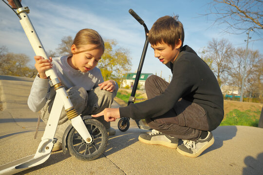 Brother pointing at a white scooter wheel helping his sister fix it on a sunny day in a park. Smart children problem solving