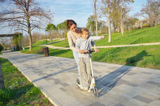Mother riding a kick scooter with her young daughter pointing forward with a fist in a sunny park. Playful authentic family active lifestyle