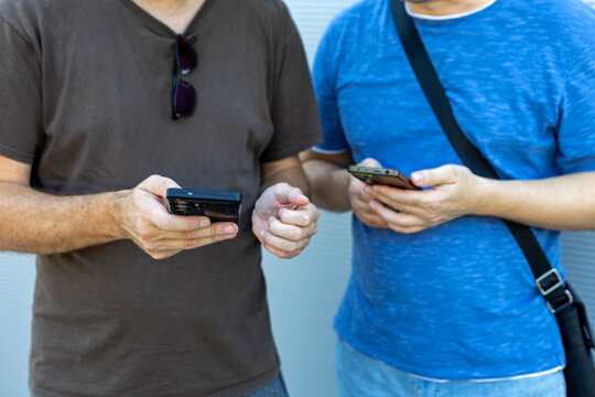Two Men Using Smartphones Side by Side. Two men standing side by side using smartphones while dressed casually against a light neutral background.
