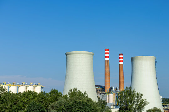 Industrial Power Plant With Cooling Towers and Smokestacks. Industrial power plant with two cooling towers, striped smokestacks, storage tanks and pipework behind green trees under a clear blue sky.