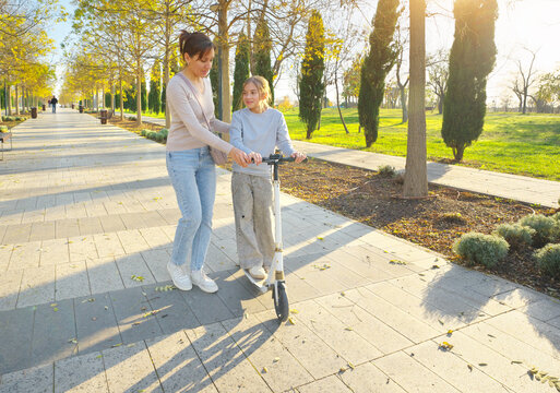 Mother guiding and helping her young daughter riding a white kick scooter along a paved park path under warm sunlight. Authentic happy family active lifestyle