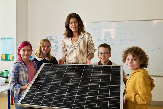 Portrait of Caucasian female teacher and diverse kids holding solar panel in classroom, learning about renewable energy. Image useful for green education campaigns