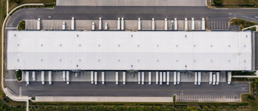 aerial top down view of large logistics warehouse with loading docks and white semi trucks parked along the building