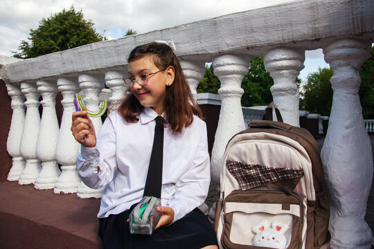 Student wearing a white shirt and tie looks happy while holding school supplies. She sits on a railing with a backpack nearby
