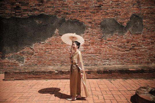 Asian woman dressed in traditional Thai outfit and umbrella with cheerful smiles at old brick wall, summer holiday travel in Ayutthaya, Southeast Asia, ancient ruins heritage,  tourist destination.