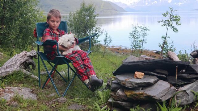 Child with pet dog, eating breakfast in the morning in front of a tent, wild camping in nature, Norway, morning, sunny morning