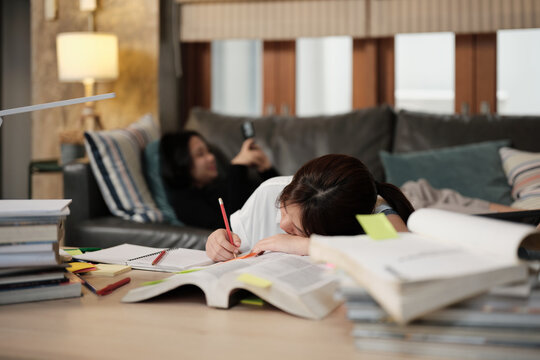 Two College Asian friends, teenage girl tired and burned out from overwhelmed homeschooling, sleeping near pile of books, stressed learning at home, her friend relaxing on sofa, education exam.