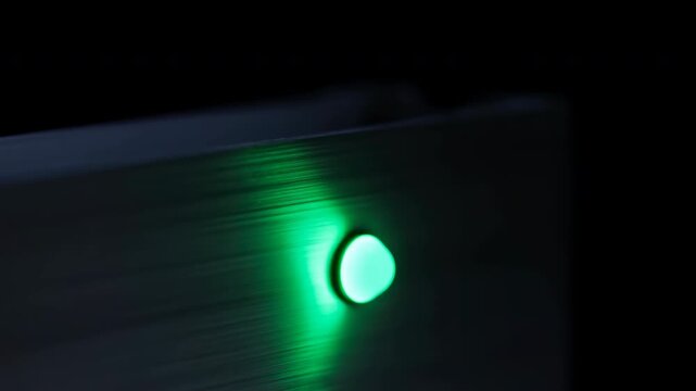 Green circular indicator light brightens on dark brushed metal control panel, casting vivid emerald glow across reflective surface in close-up low-light industrial equipment view