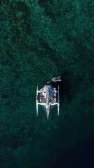 Aerial top down drone view of a white trimaran sailing yacht with a dinghy anchored on deep emerald green water above a coral reef. Overhead perspective of the boat surrounded by clear tropical sea. © Irina