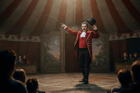 A circus ringmaster in a red tailcoat holding a hat and a whip while standing in the center of a circus tent arena before an audience