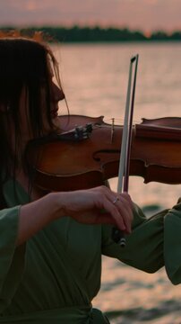 Woman playing violin by tranquil lakeside at sunset, bow gliding over strings as warm evening light reflects on water in a peaceful outdoor musical performance