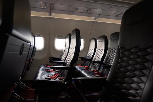 Empty black leather seats with red seatbelts line the aisle of a commercial airplane cabin. Sunlight streams through the oval windows, illuminating the modern flight interior.