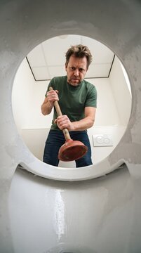 A wide-angle, point-of-view shot from inside a toilet bowl looking up at a frustrated man wearing a green t-shirt, holding a plunger and preparing to fix a clogged toilet in a white tiled bathroom.