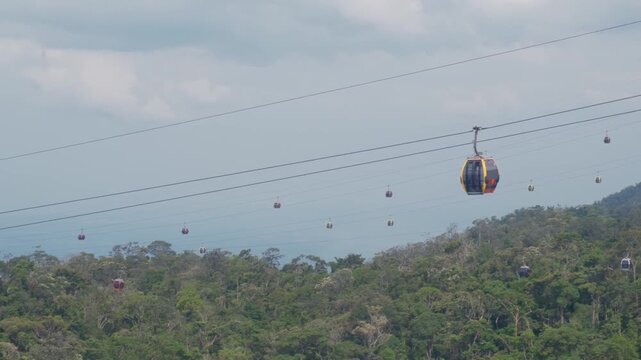 Multiple cable cars moving on suspended wires above a dense tropical green forest under a cloudy sky offering a scenic aerial view of the lush mountain landscape at Ba Na Hills Da Nang Vietnam scenery