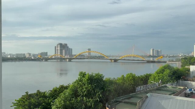 Scenic wide view of a massive yellow dragon shaped bridge spanning a calm wide river in a modern asian city skyline under a cloudy sky with green trees in the foreground and one distant bridge behind.
