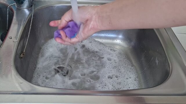 A person washes dishes in a shiny kitchen sink using soap and water. The hands scrub and rinse items under running water
