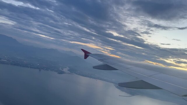 Ascending further over the coastline the passenger plane banks smoothly above the illuminated urban city lights and calm ocean waters as sunset colors fill the expansive evening sky during the ascent.