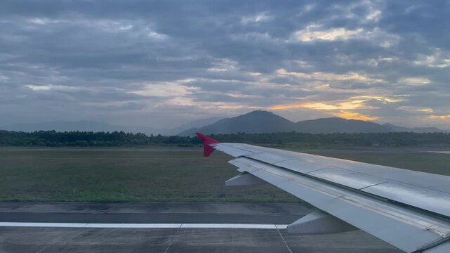Passenger window view of an airplane wing accelerating rapidly along the airport runway during sunset as the aircraft begins its departure journey with mountains and dramatic clouds in the background.