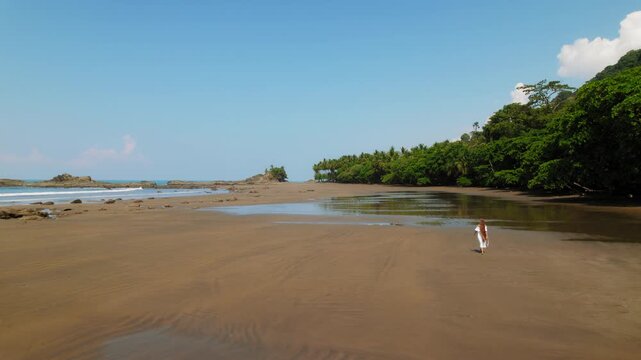 Woman walking on wide empty wet sand beach with rocks and palms on clear morning