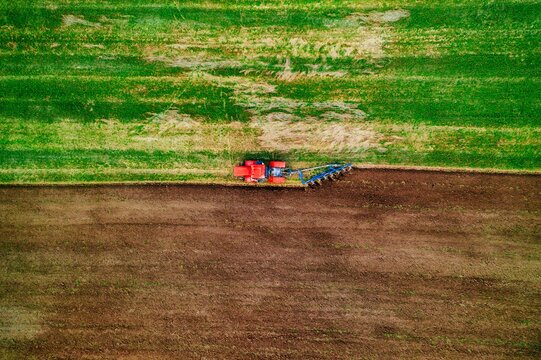Tractor preparing soil with plow, contrasting green and brown farmland from above in clear daylight