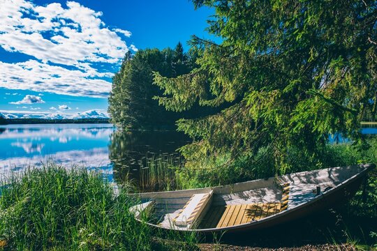 Wooden boat resting near reflective lake with lush green foliage under bright blue sky in Finland