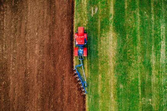 Red tractor plowing soil on brown field adjacent to vibrant green grass under clear sky