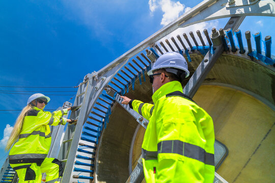 Rear three quarter view of a helmeted technician pointing along the row of stud bolts on a blade root while his colleague steadies herself on the galvanised ladder