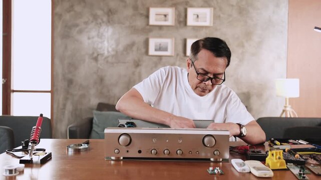 Mature Asian man disassembling and checking audio equipment, looking with magnifying glass at circuit board details for repair, home appliance DIY fix, electronics component hobby, handyman workshop.
