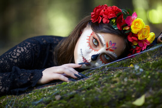 Halloween beauty portrait of young woman with Santa Muerte makeup lies on a tree trunk in the forest