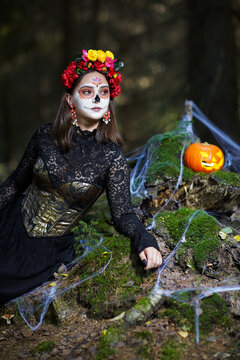 Halloween beauty portrait of young woman in the forest on a mossy stump with a halloween pumpkin.