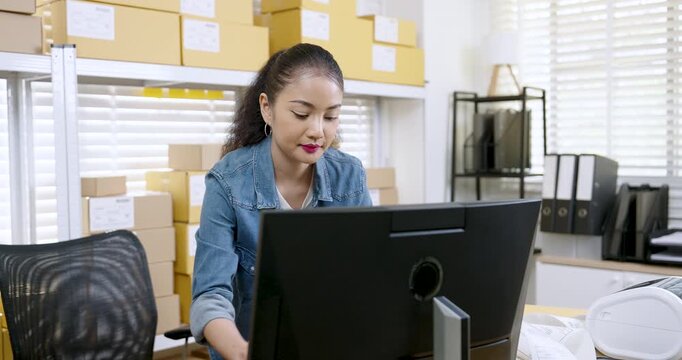 Asian woman small business owner slowly reviews customer order on computer screen, compares shipping document with online shopping parcel box list, controls e-commerce fulfillment during daily work