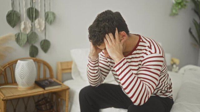 Man on bed wearing striped pyjama top, hands pressed to temples and holding face indicating headache beside bedside table with lamp in a building; anxiety reflection.