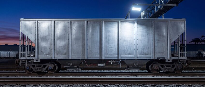 Metal hopper railcar parked on industrial tracks at night under bright overhead security lighting with other train wagons in background