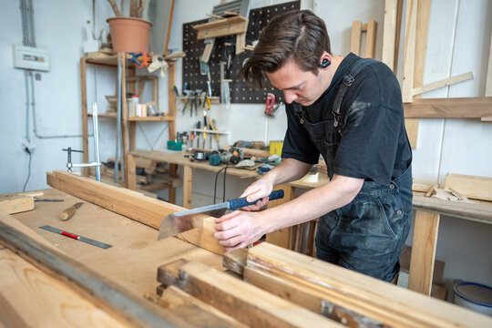 Focused restorer master preparing part of wooden frame for restoration at worktable in workshop, craftsman using hand saw instrument to remove damaged part, carefully cutting before patch installing.