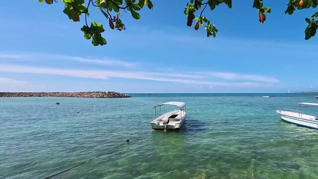 Two small motorboats float on turquoise water near shore, framed by leaves, with a stone breakwater and bright sky in the background. Ideal for tropical travel and beach vacation concepts.