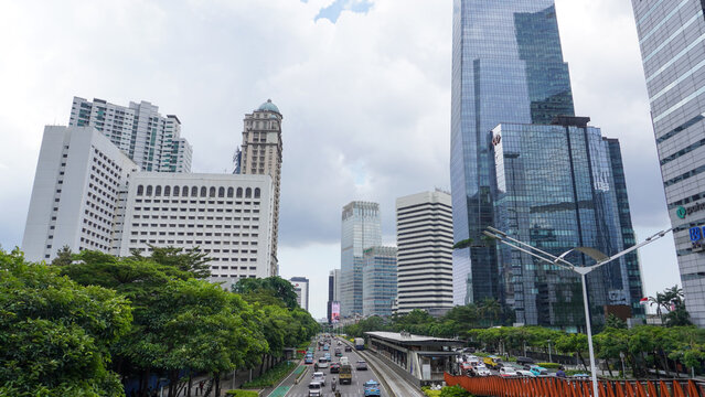 Bustling traffic on Sudirman street with modern skyscrapers, lush green trees, and elevated infrastructure in Karet district, Jakarta, Indonesia under a cloudy day sky. Wide angle cityscape view.