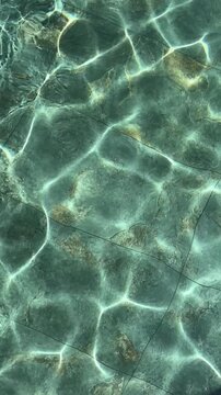 Clear turquoise water ripple with sun caustics on natural stone tile pool bottom for summer and spa concept