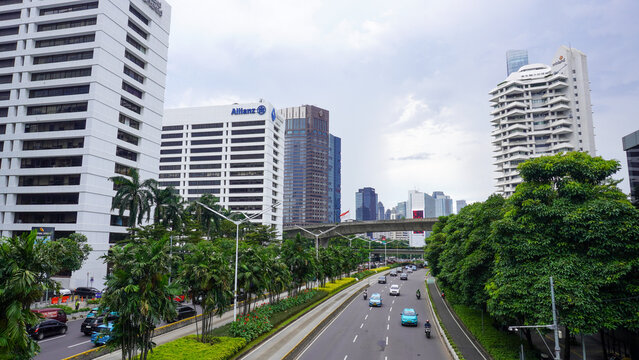 Bustling traffic on Sudirman street with modern skyscrapers, lush green trees, and elevated infrastructure in Karet district, Jakarta, Indonesia under a cloudy day sky. Wide angle cityscape view.