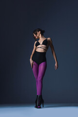 Fototapeta premium Full length image of a sexy, beautiful dancer dressed in a sensual outfit with purple tights, moving in dance poses in the studio, against a dark background.