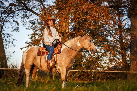 Woman in cowboy hat sitting on horse enjoying western lifestyle. Cowgirl horseback riding in autumn nature during sunset