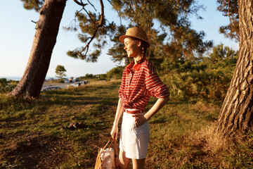 Naklejka premium Young woman enjoying lifestyle outdoor nature portrait. She wears straw hat and striped shirt standing in park forest at sunset. Summer mood concept for travel and vacation.