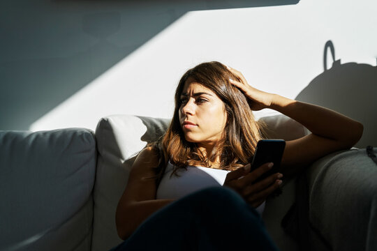 Young white woman holding mobile device sitting thoughtfully on couch