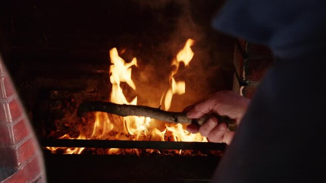 Person stirring embers with a wooden stick inside a brick fireplace to maintain fire.