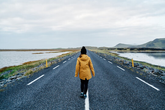 A white woman walks on an empty road surrounded by water and hills