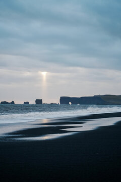 A sunbeam shines through clouds over black sand beach and ocean rocks