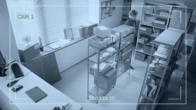 Young Caucasian man wearing headphones entering storage room, checking phone, and typing on laptop, filmed from high angle CCTV camera. Footage suiting security, logistics, or loss prevention promos